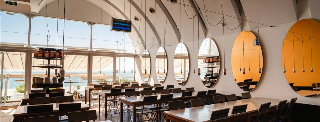 Salão amplo de refeições do restaurante Casanova com mesas compridas, espelhos ovais decorativos e vista através de grandes janelas de vidro.