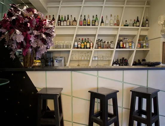 Balcão de bar do restaurante Casa Nostra com bancos altos de madeira, estante geométrica com garrafas e arranjo decorativo floral.