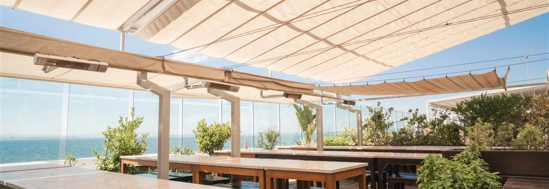 Esplanada exterior coberta do restaurante Casanova com mesas compridas, plantas e vista panorâmica para as águas do rio.