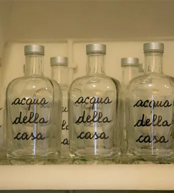 Garrafas de vidro reutilizáveis com inscrição Acqua della Casa alinhadas na prateleira do restaurante Casa Nostra.
