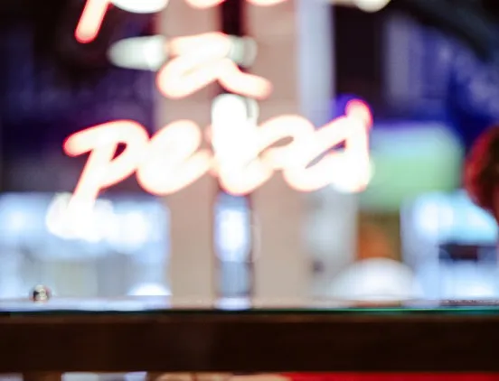 Letreiro luminoso em néon avermelhado ao fundo do balcão da pizzaria Pizza a Pezzi.