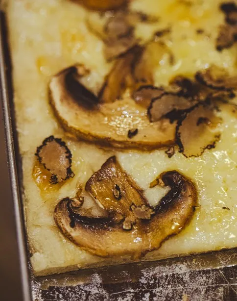 Fatias de cogumelos salteados sobre base de massa de pizza retangular no tabuleiro da Pizza a Pezzi.