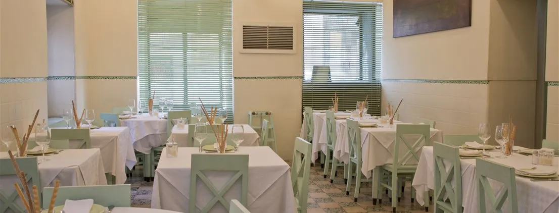Salão de refeições do restaurante Casa Nostra com mesas postas com toalhas brancas, cadeiras verde-claras e janelas com persianas.